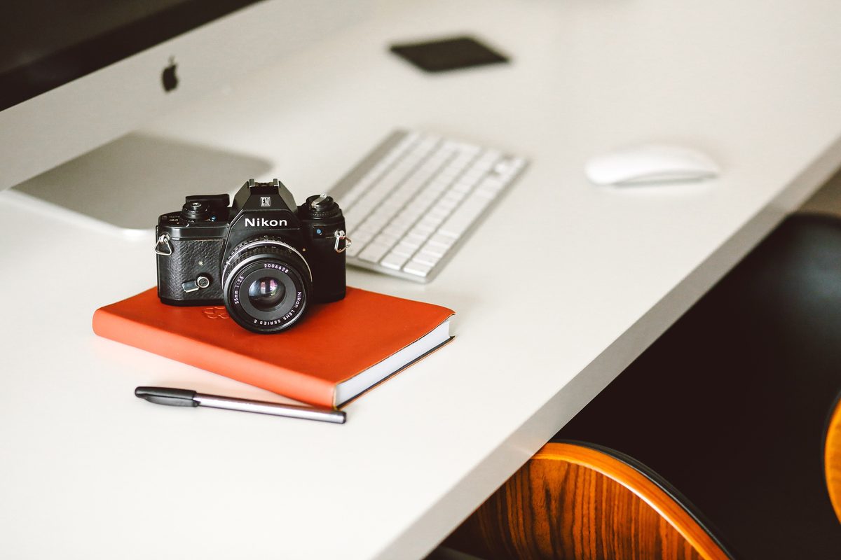 Aparat fotograficzny Nikon leży na pomarańczowym notesie na białym biurku, obok długopis, klawiatura i myszka komputera