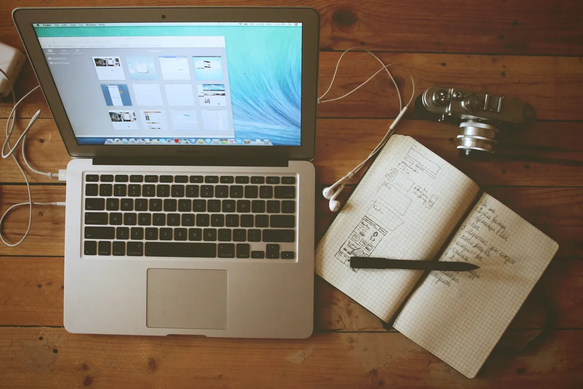 JPEG XL a Grafika Google i Discover: nowy format zdjęć tylko z wersją zapasową Otwarty laptop na drewnianym stole z biblioteką zdjęć na ekranie, obok aparat fotograficzny, słuchawki i zeszyt w kratkę ze szkicami